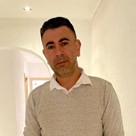 Fachmann für Objektbetreuung Ein Mann mit kurzen schwarzen Haaren trägt einen grauen Pullover in einem hellen Raum.