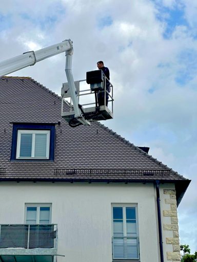 Dachrinnen- und Dachreinigung Arbeiter auf einer Hebebühne repariert das Dach eines Hauses unter einem wolkigen Himmel.